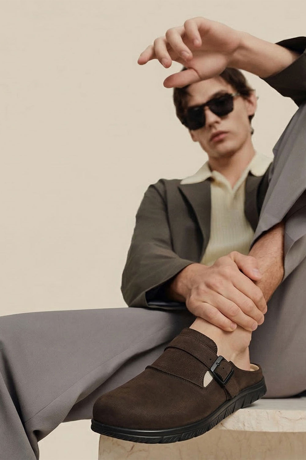 Man wearing sunglasses and a gray suit with brown shoes on a beige background